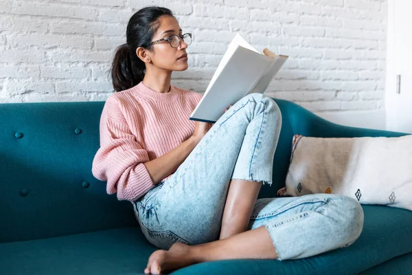 Bella giovane donna che legge un libro mentre è seduta sul divano a casa . — Foto Stock