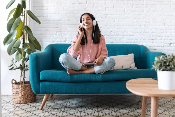 Mujer bastante joven escuchando música con tableta digital y relajándose mientras se sienta en el sofá en casa . — Foto de Stock