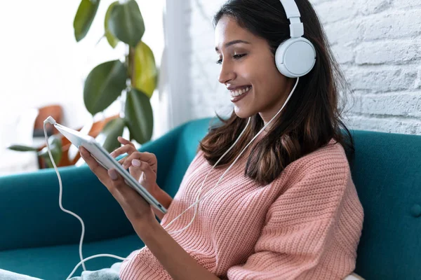 Hübsche junge Frau hört Musik mit digitalem Tablet, während sie zu Hause auf dem Sofa sitzt. — Stockfoto