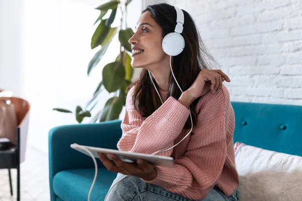Hübsche junge Frau hört Musik mit digitalem Tablet, während sie zu Hause auf dem Sofa sitzt. — Stockfoto