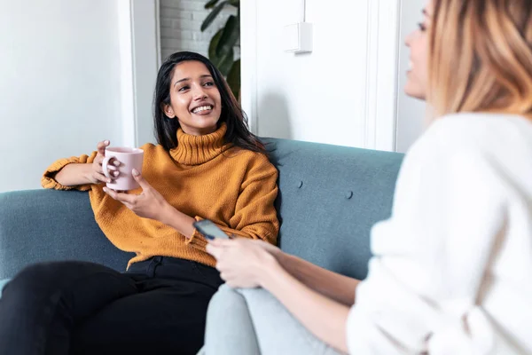 Dos mujeres jóvenes y bonitas usando el teléfono móvil mientras beben café en el sofá en casa . — Foto de Stock