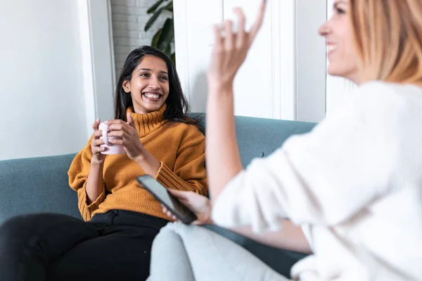 Deux jolies jeunes femmes utilisant un téléphone portable tout en buvant du café sur le canapé à la maison . — Photo