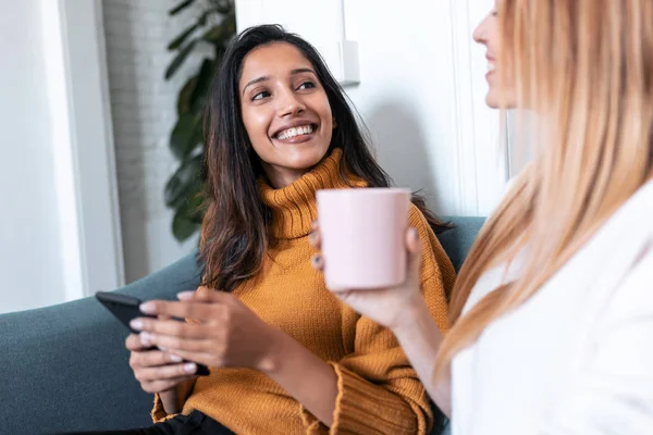 Dos mujeres jóvenes y bonitas usando el teléfono móvil mientras beben café en el sofá en casa . — Foto de Stock