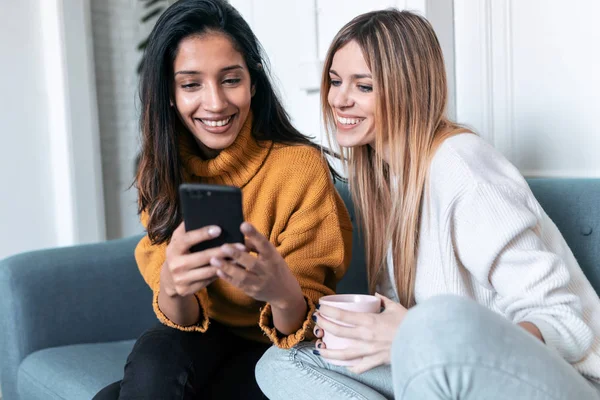 Duas jovens mulheres bonitas usando telefone celular enquanto bebem café no sofá em casa . — Fotografia de Stock