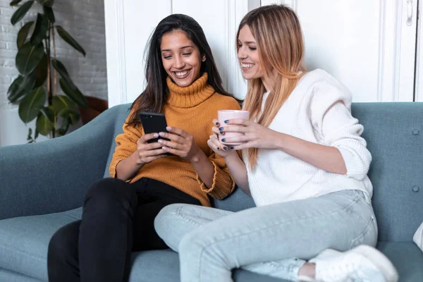 Dos mujeres jóvenes y bonitas usando el teléfono móvil mientras beben café en el sofá en casa . — Foto de Stock