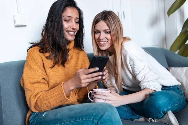 Dos mujeres jóvenes y bonitas usando el teléfono móvil mientras beben café en el sofá en casa . — Foto de Stock