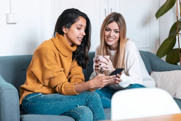 Twee mooie jonge vrouwen met behulp van mobiele telefoon tijdens het drinken van koffie op de bank thuis. — Stockfoto