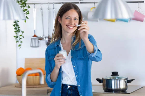 Mulher bonita olhando para a câmera enquanto come iogurte na cozinha em casa . — Fotografia de Stock
