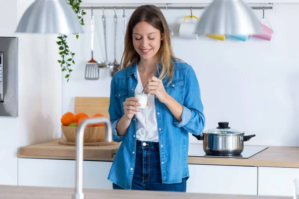 Красивая молодая женщина ест йогурт, стоя дома на кухне . — стоковое фото