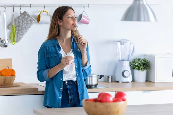 Красивая молодая женщина ест йогурт, стоя дома на кухне . — стоковое фото