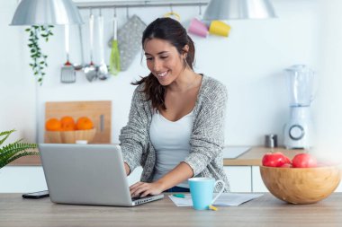 Evde mutfakta dizüstü bilgisayar ve belgelerle çalışan güzel genç kadın.