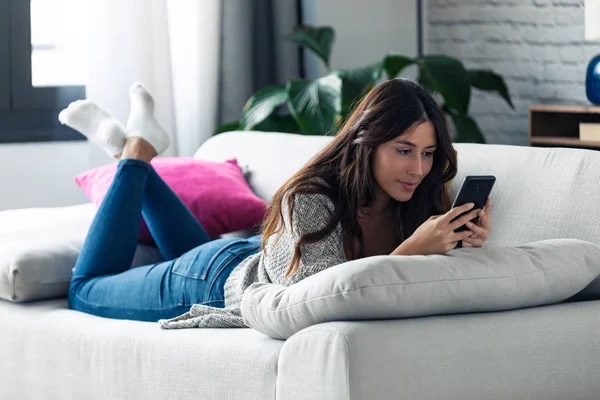 Söt ung kvinna skickar meddelanden med sin mobiltelefon när hon ligger på soffan hemma. — Stockfoto