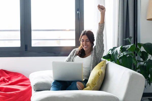 Mujer bastante joven celebrando algo mientras usa su computadora portátil en el sofá en casa . —  Fotos de Stock