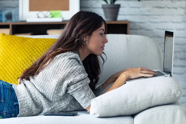Söt ung kvinna som använder sin bärbara dator när hon ligger på soffan hemma. — Stockfoto