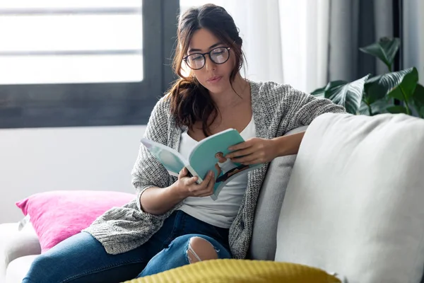 Mujer bastante joven con anteojos leyendo un libro mientras está sentada en el sofá en casa . —  Fotos de Stock
