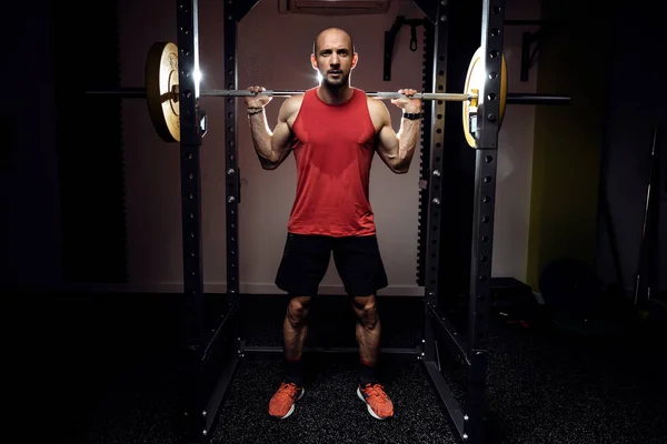 Sterke gespierde bodybuilder atletische man pompen spieren met lange halter op donkere studio. — Stockfoto
