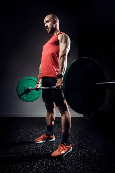 Strong muscular bodybuilder athletic man pumping up muscles with barbell on dark studio. — Stockfoto