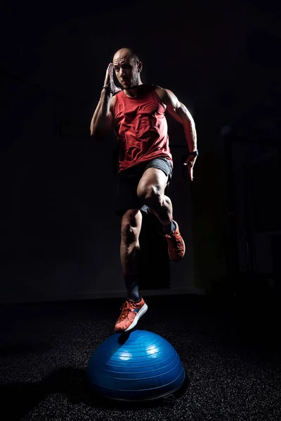 Fit atleet uitvoeren van oefening op gymnastisch halfrond boezem bal op donkere studio. — Stockfoto