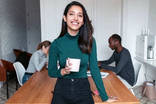 Giovane imprenditrice indiana che guarda la telecamera in ufficio. Sullo sfondo, i suoi colleghi che lavorano . — Foto Stock