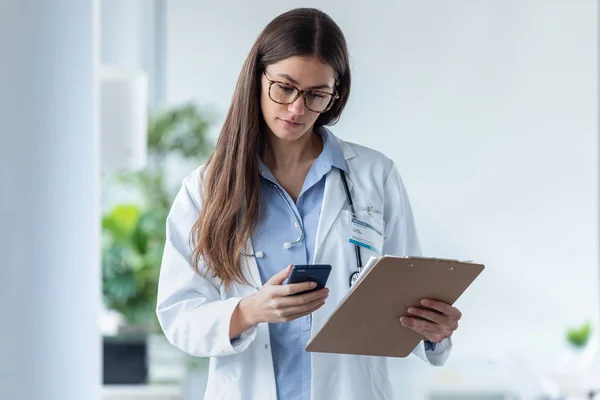 Medico piuttosto femminile che utilizza il suo telefono cellulare durante la revisione dei rapporti medici in consultazione medica . — Foto Stock