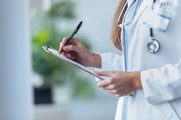 Kobieta lekarz wypełnienie formularza medycznego w schowku podczas stojąc w konsultacji. — Zdjęcie stockowe