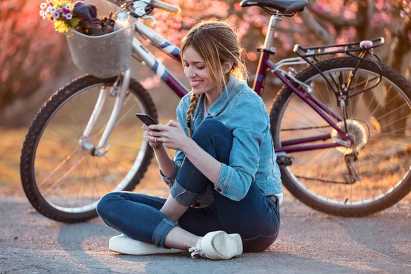 Jolie jeune femme avec un vélo vintage utilisant son téléphone portable sur le champ de cerises au printemps . — Photo