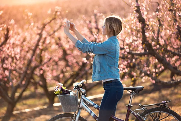 Pretty young woman with a vintage bike taking photographs of cherry blossoms on the field in springtime. — 스톡 사진