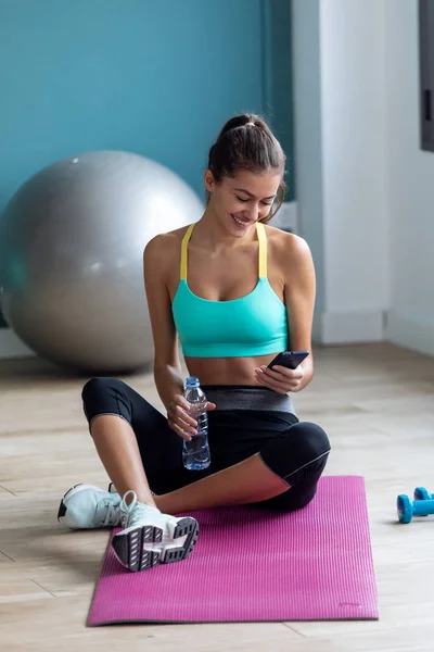 Sportowa młoda kobieta używa telefonu komórkowego trzymając butelkę wody po zajęciach pilatesu na siłowni. — Zdjęcie stockowe