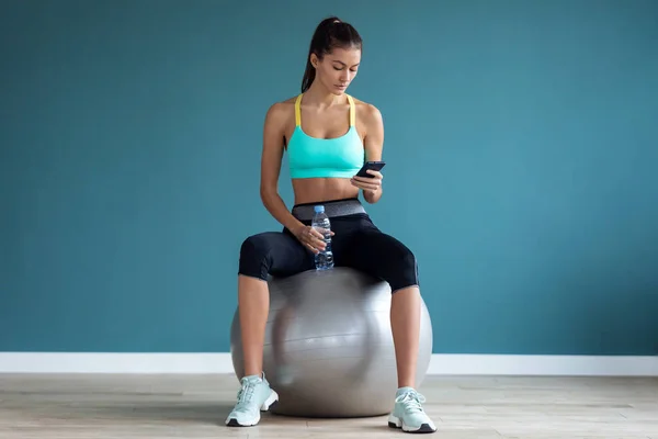 Jeune femme sportive utilisant son téléphone portable tout en tenant une bouteille d'eau après un cours de pilates sur la salle de gym . — Photo