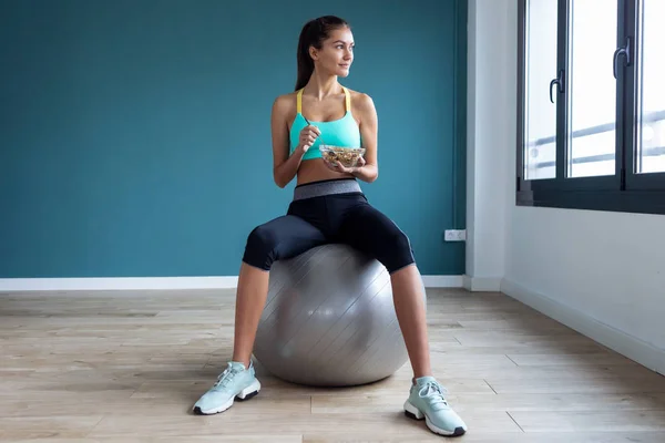 Sporty young woman eating a bowl of muesli while sitting on fitness ball at home.