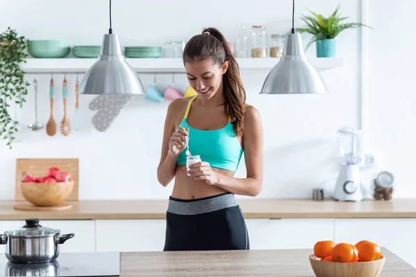 Deportiva mujer joven comiendo yogur mientras está de pie en la cocina en casa . — Foto de Stock