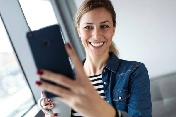 Evdeki oturma odasının penceresinin yanında kahve içerken selfie çeken güzel genç bir kadın.. — Stok fotoğraf