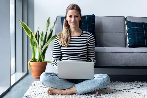 Mujer bastante joven mirando a la cámara mientras usa su computadora portátil en casa . —  Fotos de Stock