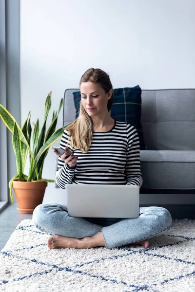 Mujer bastante joven usando su teléfono móvil mientras trabaja con el ordenador portátil sentado en el suelo en casa . —  Fotos de Stock