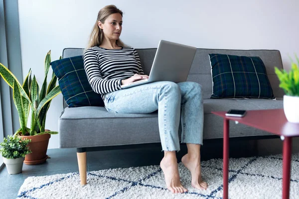 Mujer bastante joven usando su computadora portátil mientras está sentada en el sofá en casa . —  Fotos de Stock