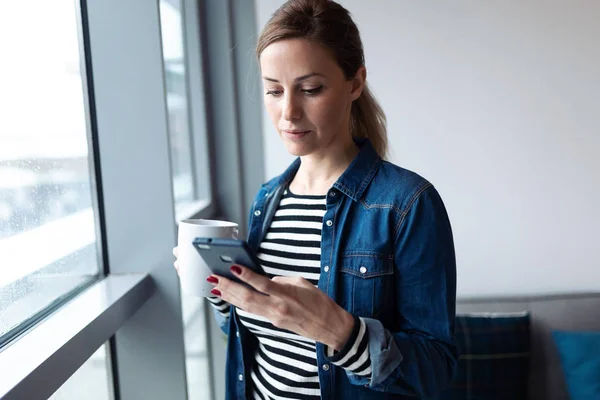 Hübsche junge Frau mit ihrem Handy beim Kaffeetrinken am Fenster im heimischen Wohnzimmer. — Stockfoto