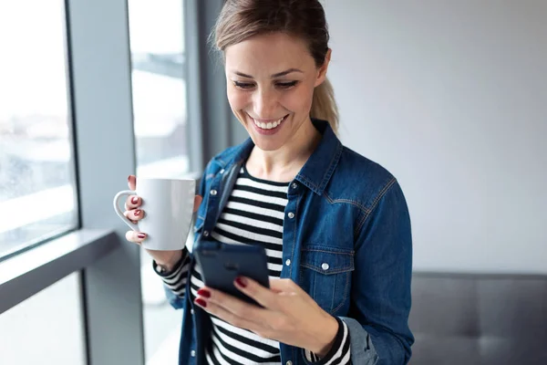 Hezká mladá žena pomocí mobilního telefonu při pití kávy v blízkosti okna v obývacím pokoji doma. — Stock fotografie