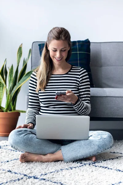 Mujer bastante joven usando su teléfono móvil mientras trabaja con el ordenador portátil sentado en el suelo en casa . —  Fotos de Stock