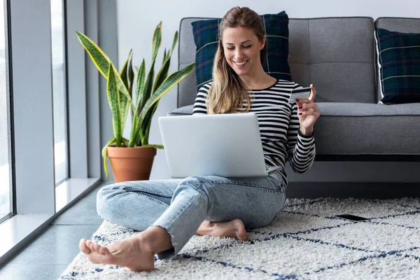 Söt ung kvinna shopping online med kreditkort och bärbar dator när du sitter på golvet hemma. — Stockfoto