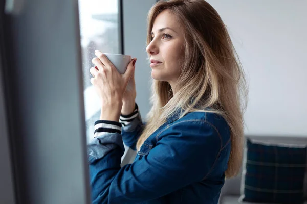 Mujer bastante joven mirando a través de la ventana mientras bebe café en la sala de estar en casa . — Foto de Stock