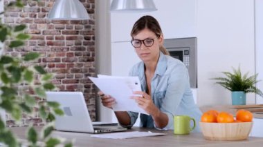 Evde mutfakta dizüstü bilgisayar ve belgelerle çalışan güzel genç kadın.