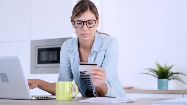 Όμορφη νεαρή γυναίκα που χρησιμοποιεί το laptop της για να ψωνίσει στο διαδίκτυο και να πληρώσει με πιστωτική κάρτα ενώ κάθεται στην κουζίνα στο σπίτι. — Αρχείο Βίντεο
