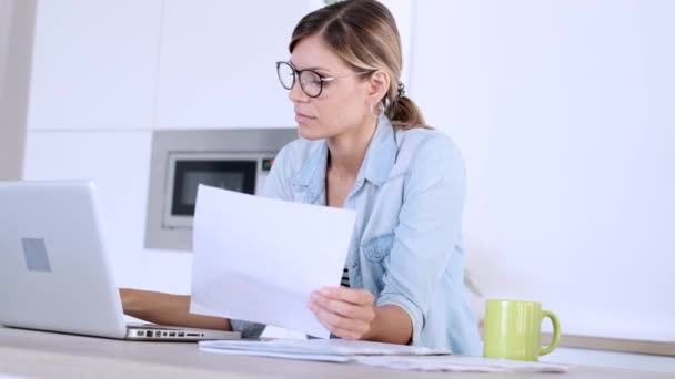 Jolie jeune femme travaillant avec un ordinateur portable et des documents dans la cuisine à la maison . — Video