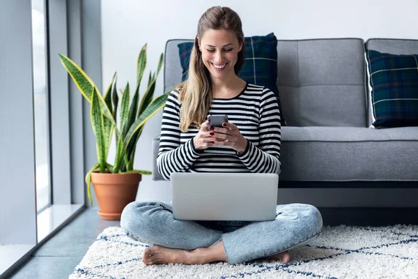 Wanita muda cantik menggunakan ponselnya saat bekerja dengan laptop duduk di lantai di rumah . — Stok Foto