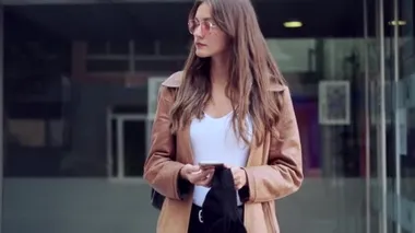 Video of pretty young woman looking at camera while holding mobile phone and standing in the street.