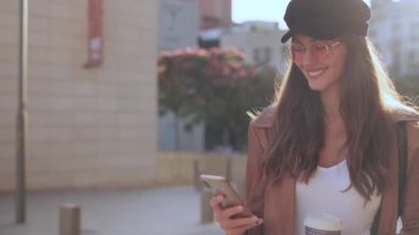 Video of pretty young woman using her mobile phone while holding a coffee and standing in the street.