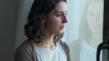 Video of sad young woman crying while looking through the window at home.