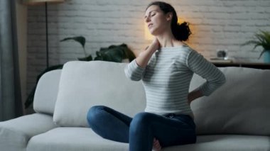 Video of young woman with back and neck pain sitting on the sofa in the living room at home.