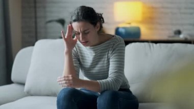 Video of young woman with headache sitting on the sofa in the living room at home.