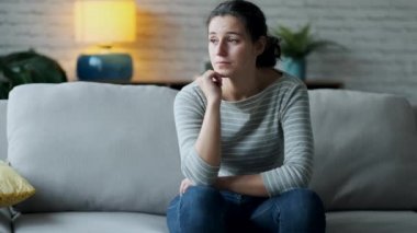 Video of depressed young woman thinking about her problems while sitting on the sofa at home.
